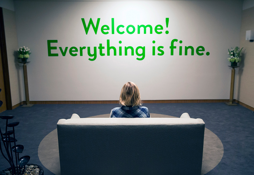 Woman sitting on a couch reading words on the wall saying, “Welcome! Everything is fine”.
